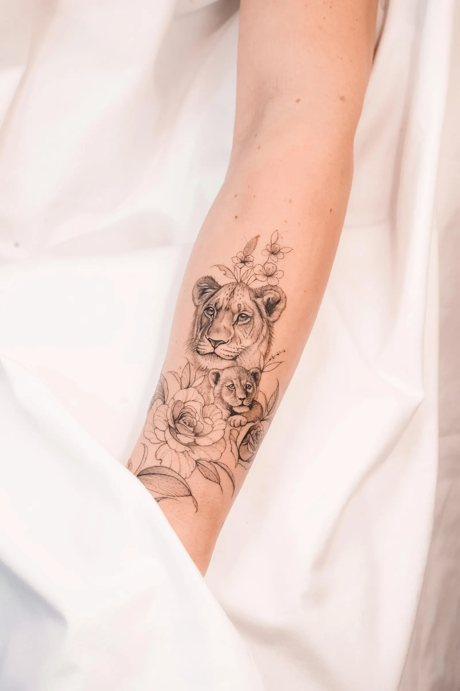 Lioness and cub with roses and leaves, tattooed along the forearm on white fabric.