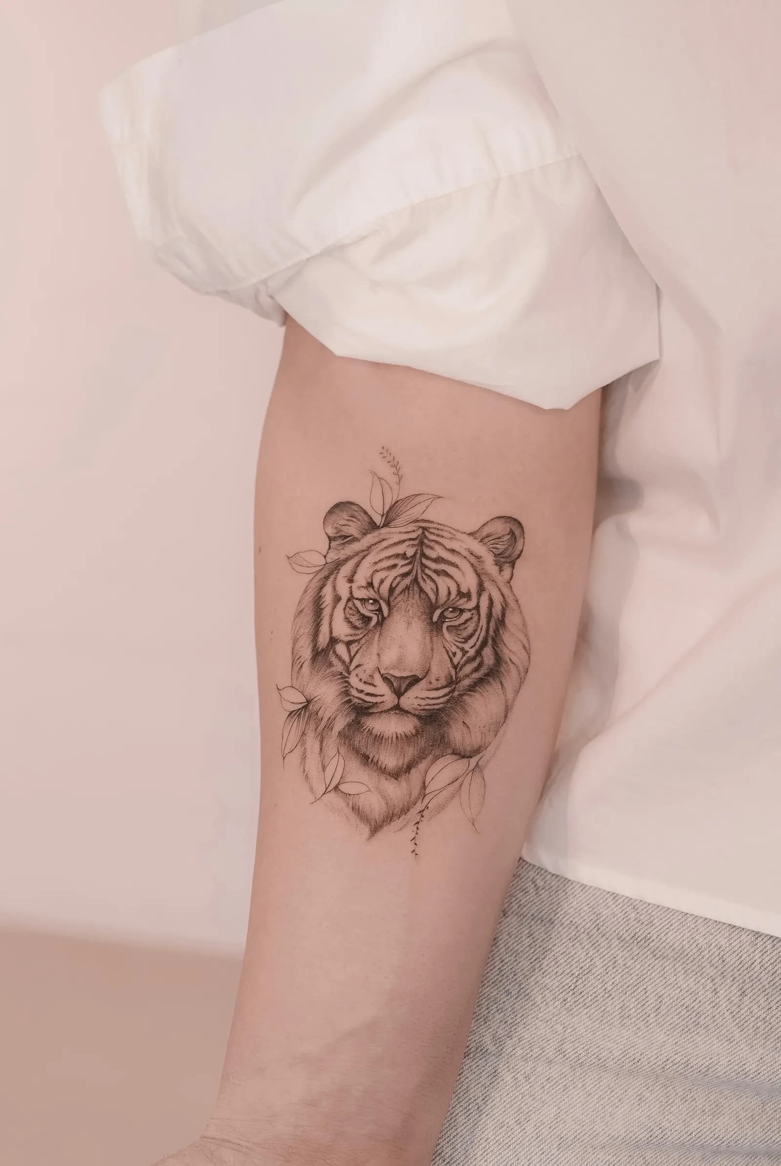 Black-and-grey tiger head framed by leaves tattooed on a forearm.