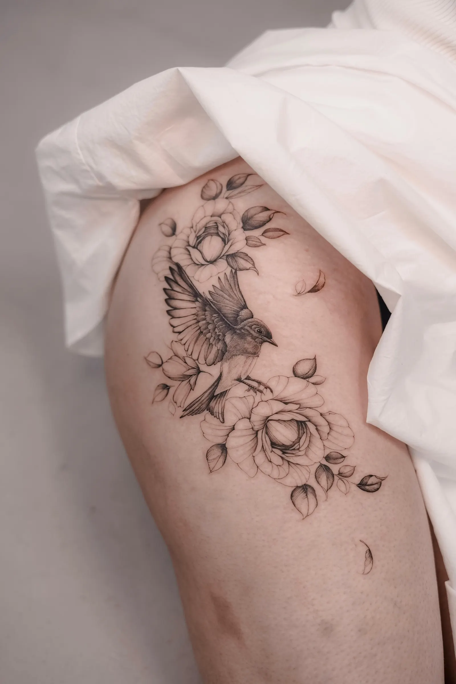Bird in flight among peonies and falling petals, tattooed across the hip and thigh.