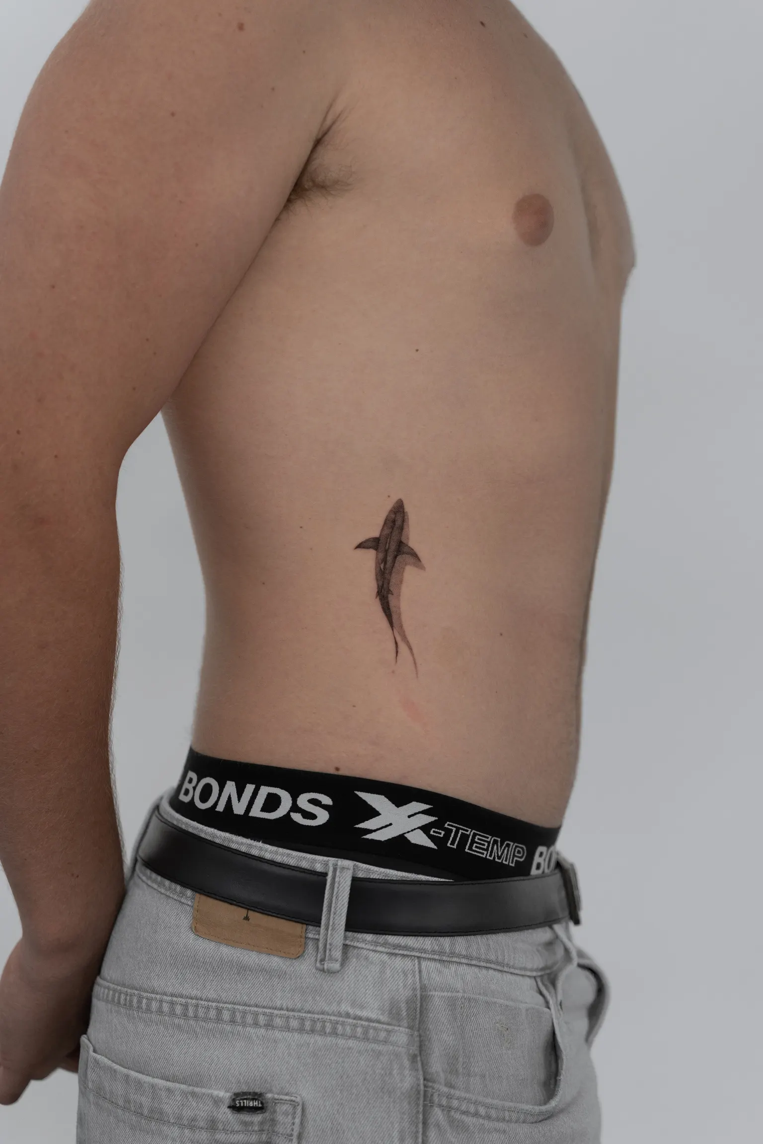 Small shaded shark tattoo on the side of the torso above the waistband.