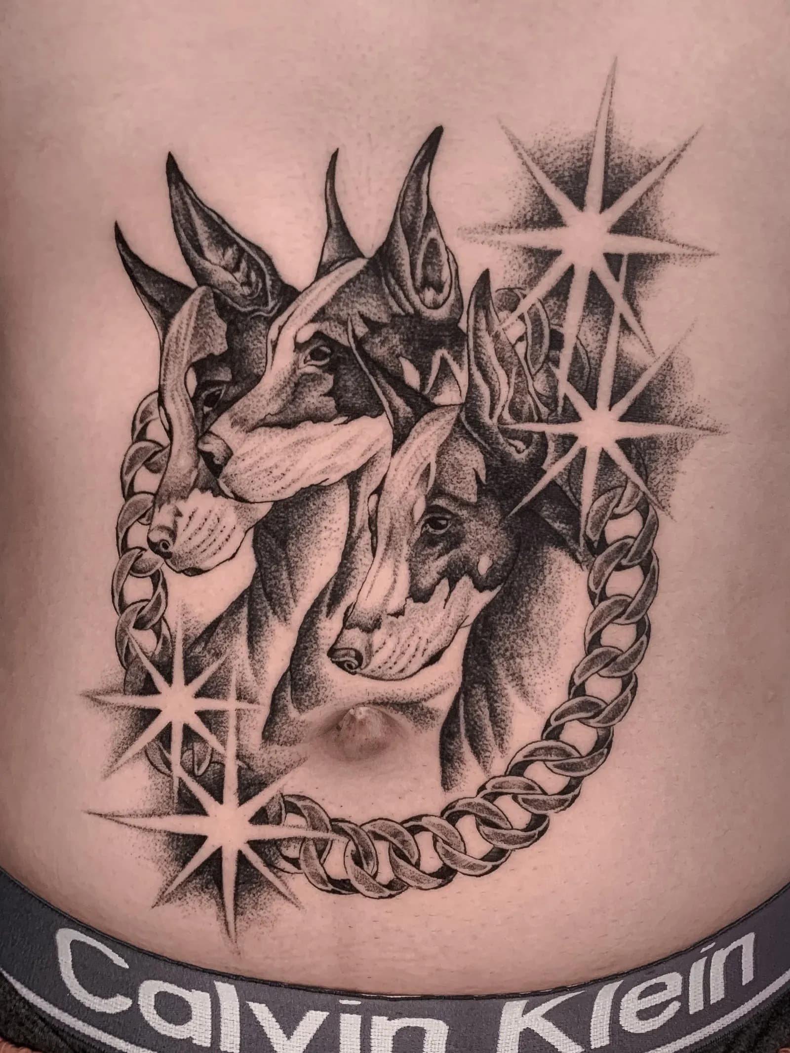 Black and grey tattoo of three doberman heads with a chain and stars on a stomach.