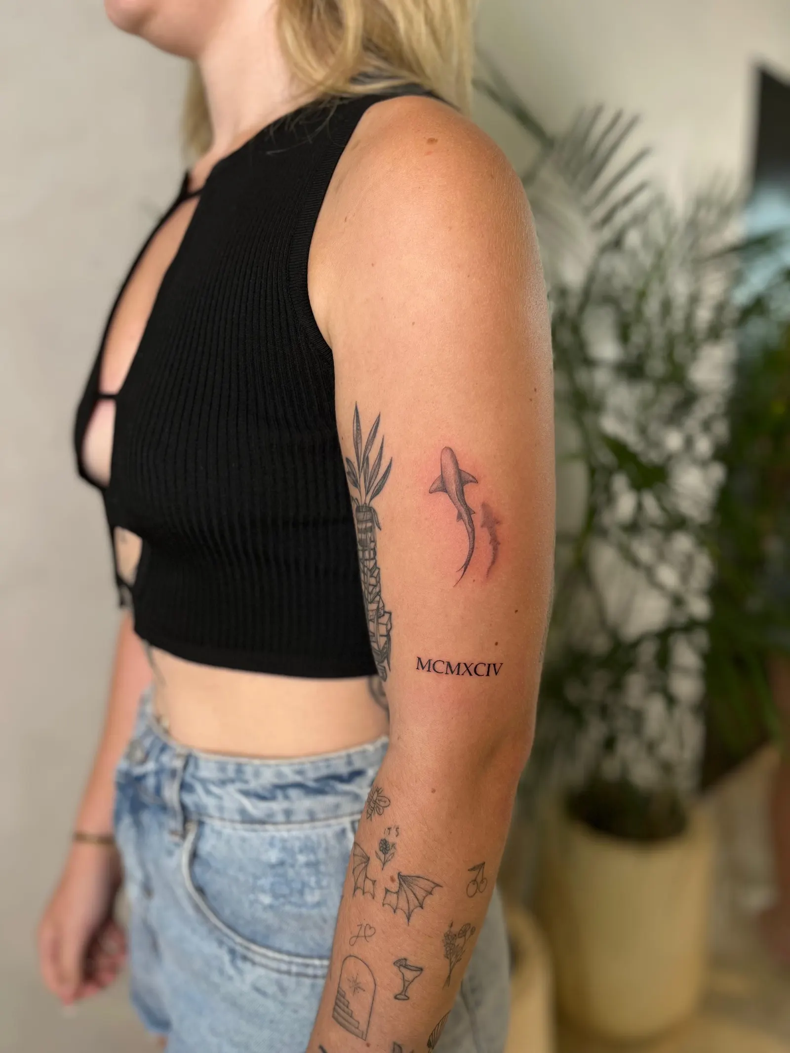 Small black-and-grey shark above Roman numerals tattooed on an upper arm.