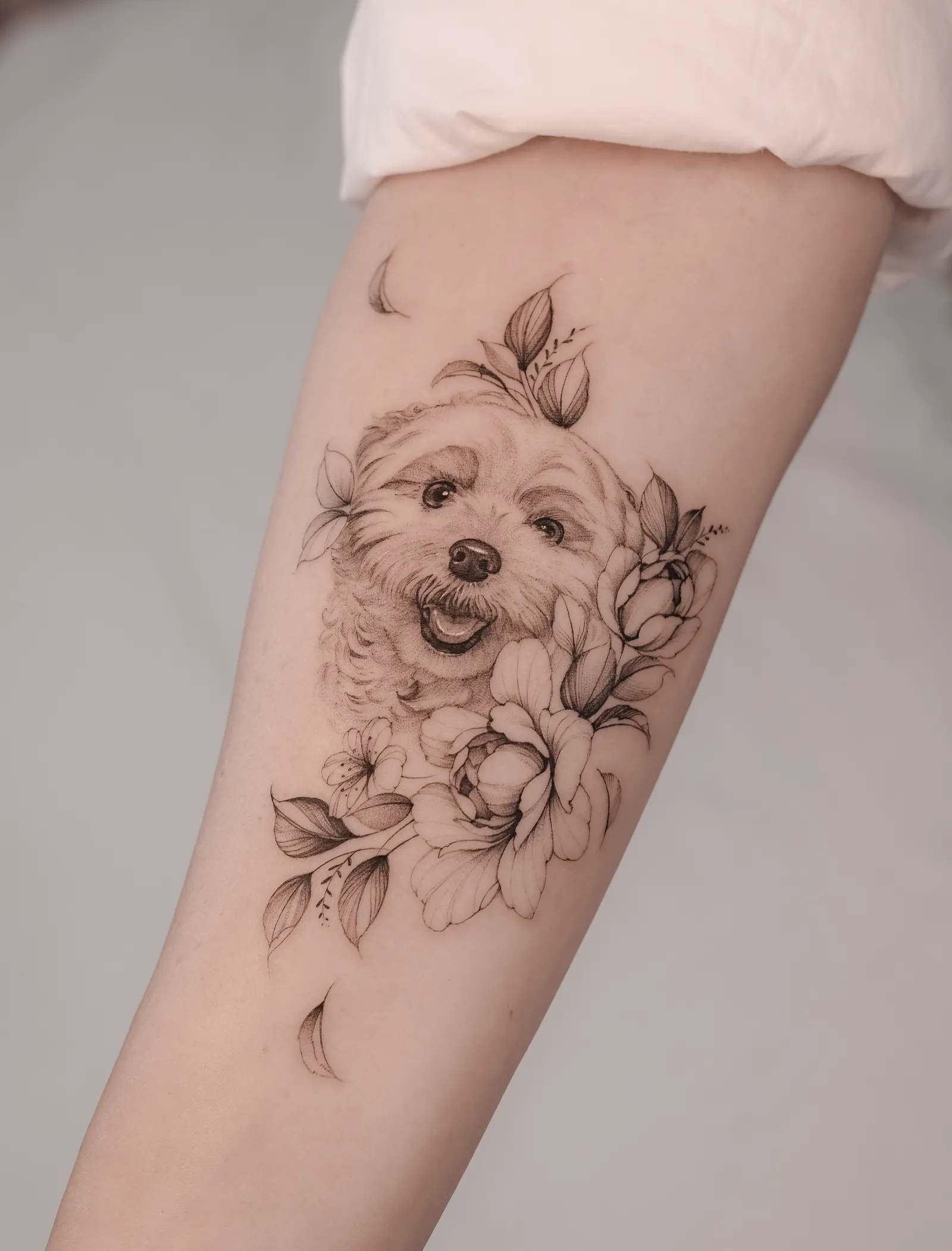 Portrait of a small fluffy dog surrounded by peonies and leaves, tattooed on the arm.