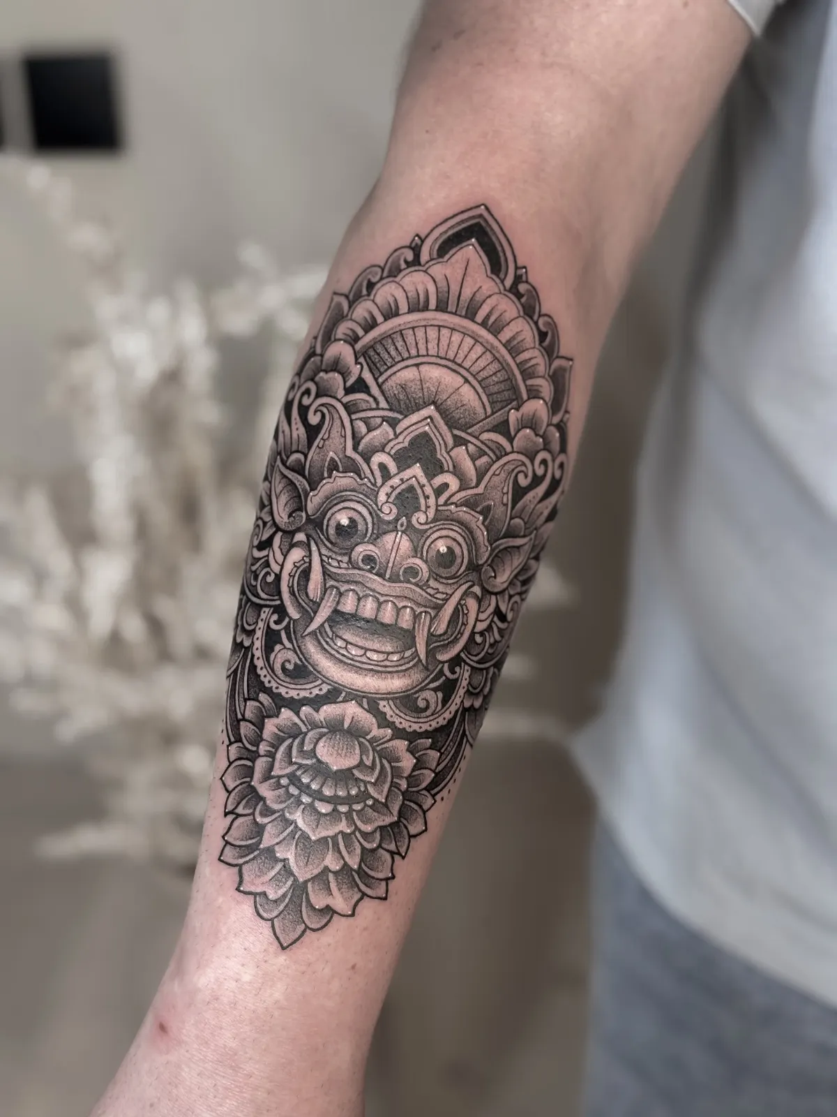 Ornamental mask tattoo with lotus flowers in black and grey on a forearm.