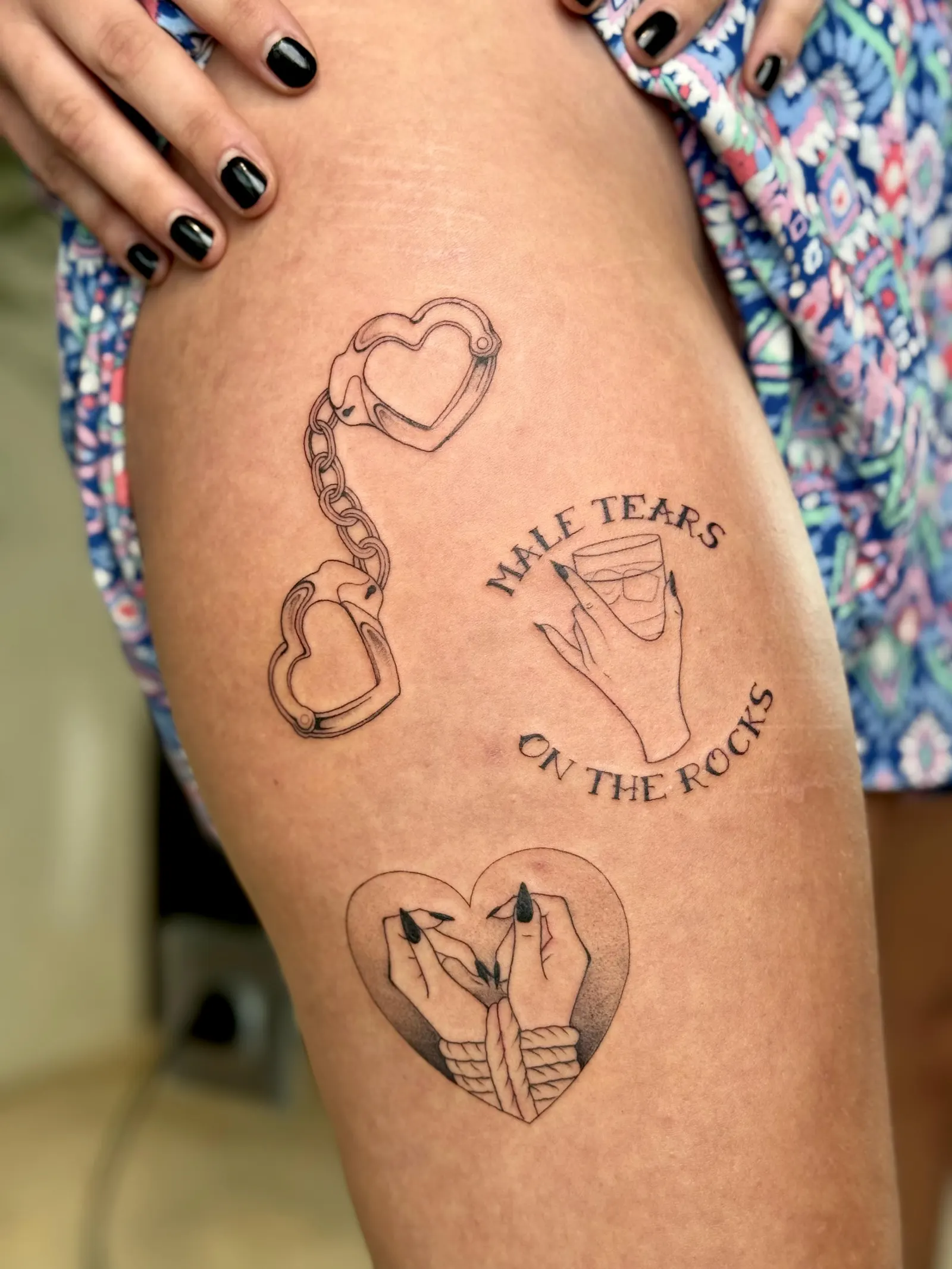 Three fine-line thigh tattoos: heart handcuffs, a hand holding a glass with text, and clasped hands.