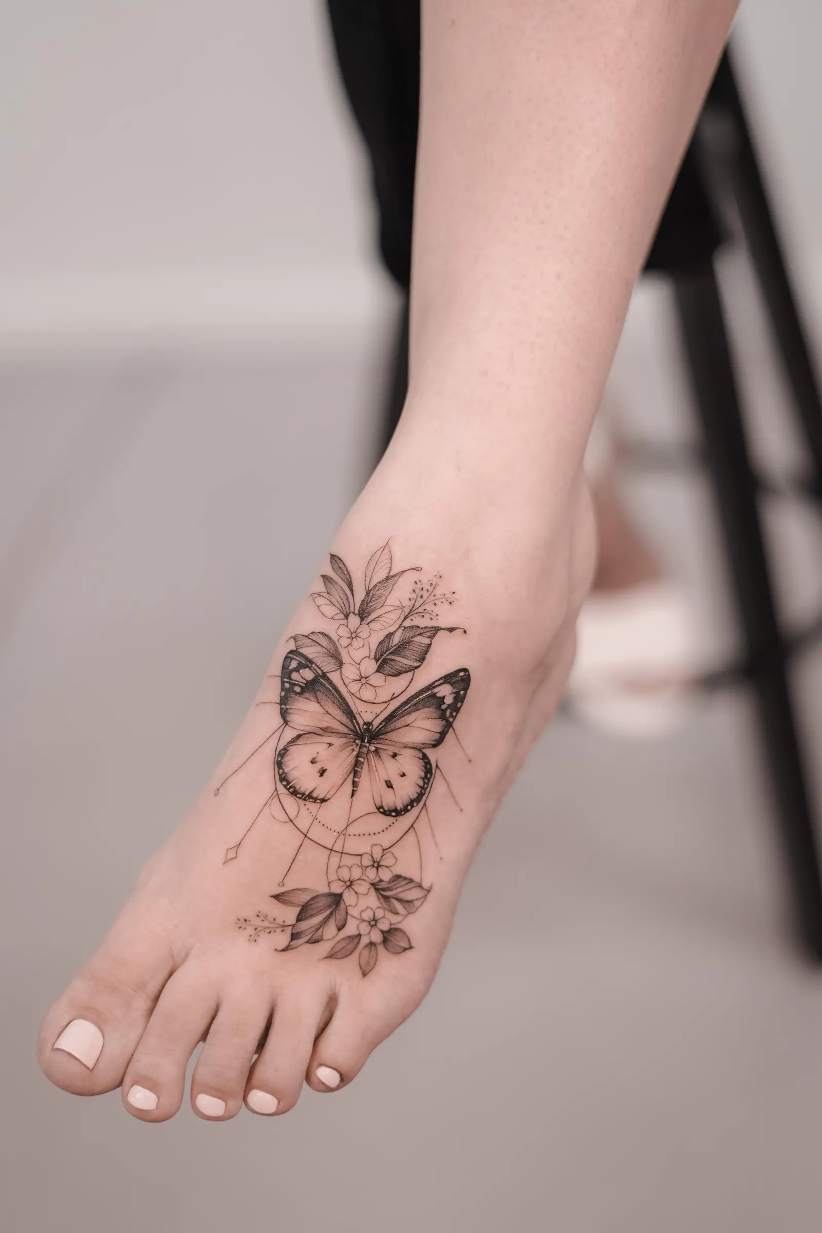 Butterfly with flowers and fine geometric lines tattooed on the top of the foot.