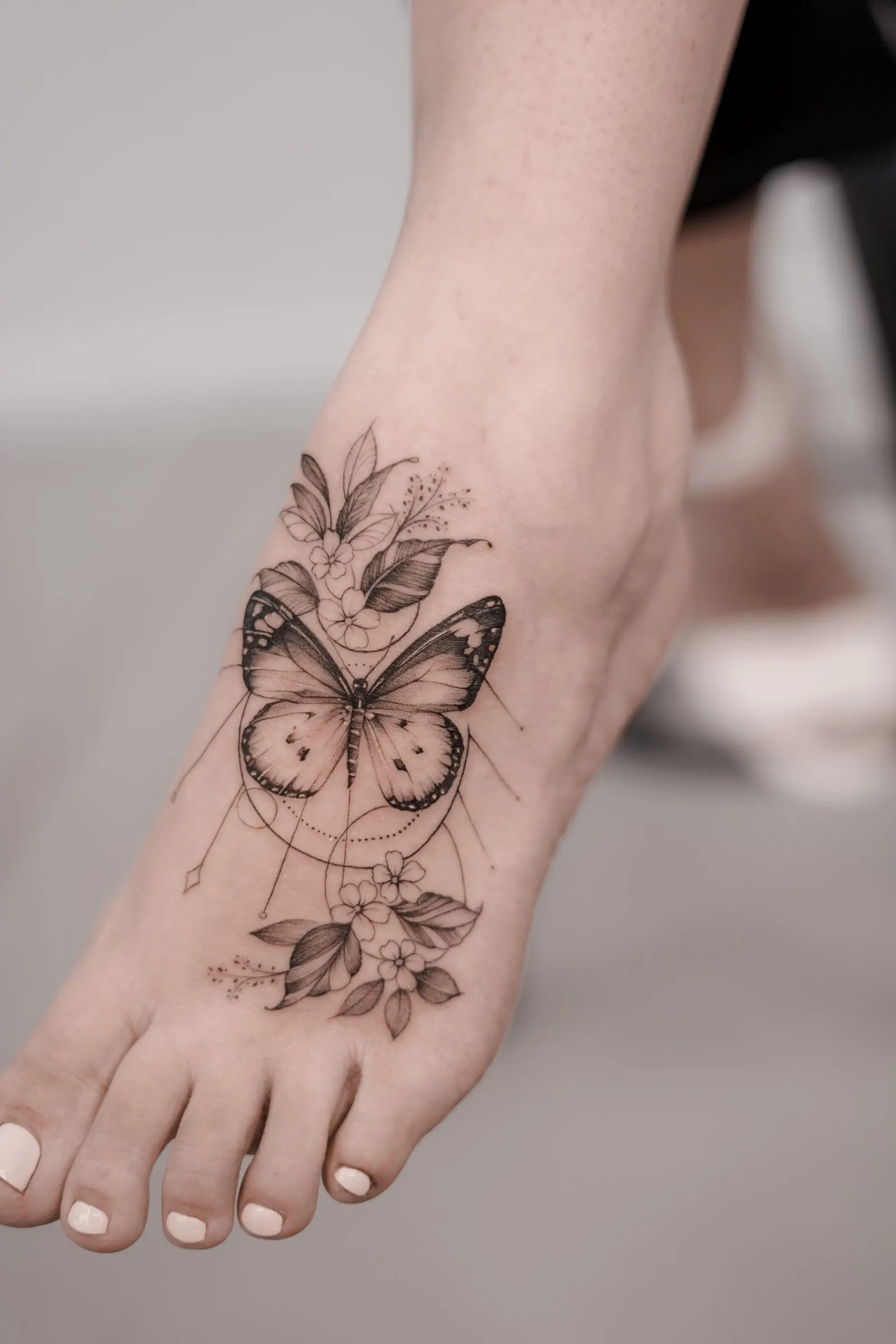 Black and grey butterfly tattoo with flowers and thin geometric lines on a foot.