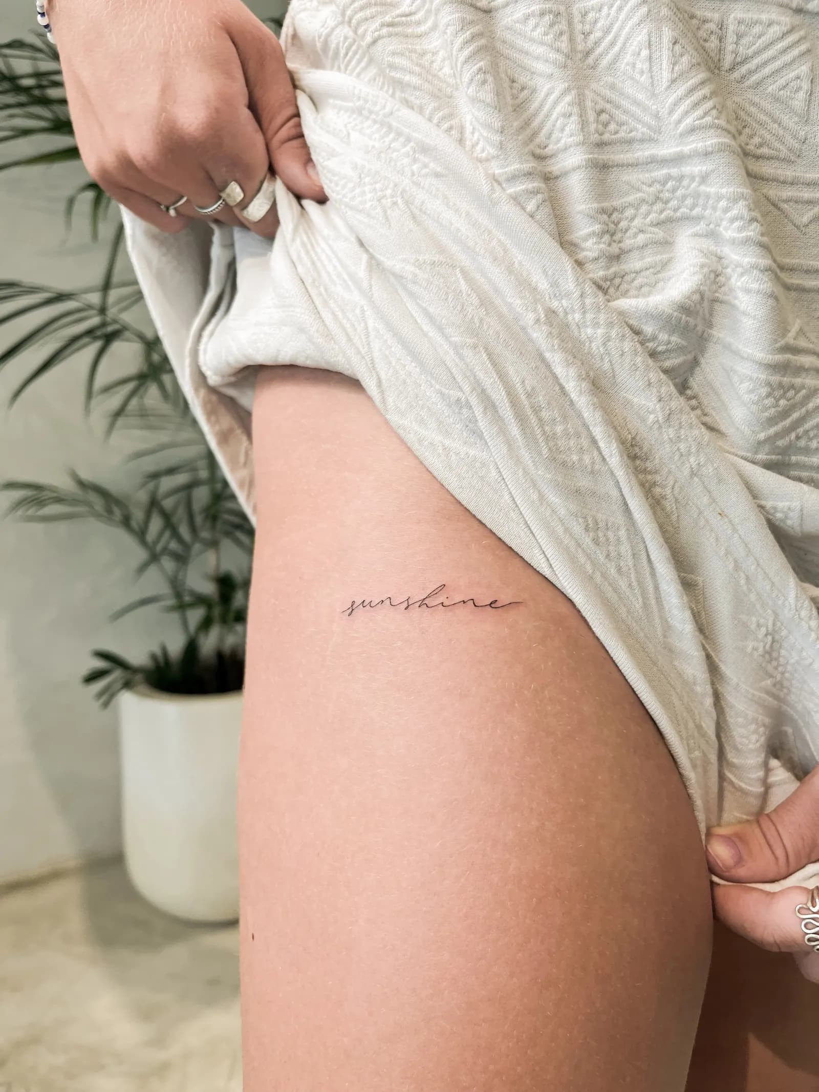 Fine script word tattoo on an outer thigh with a knit top held aside.