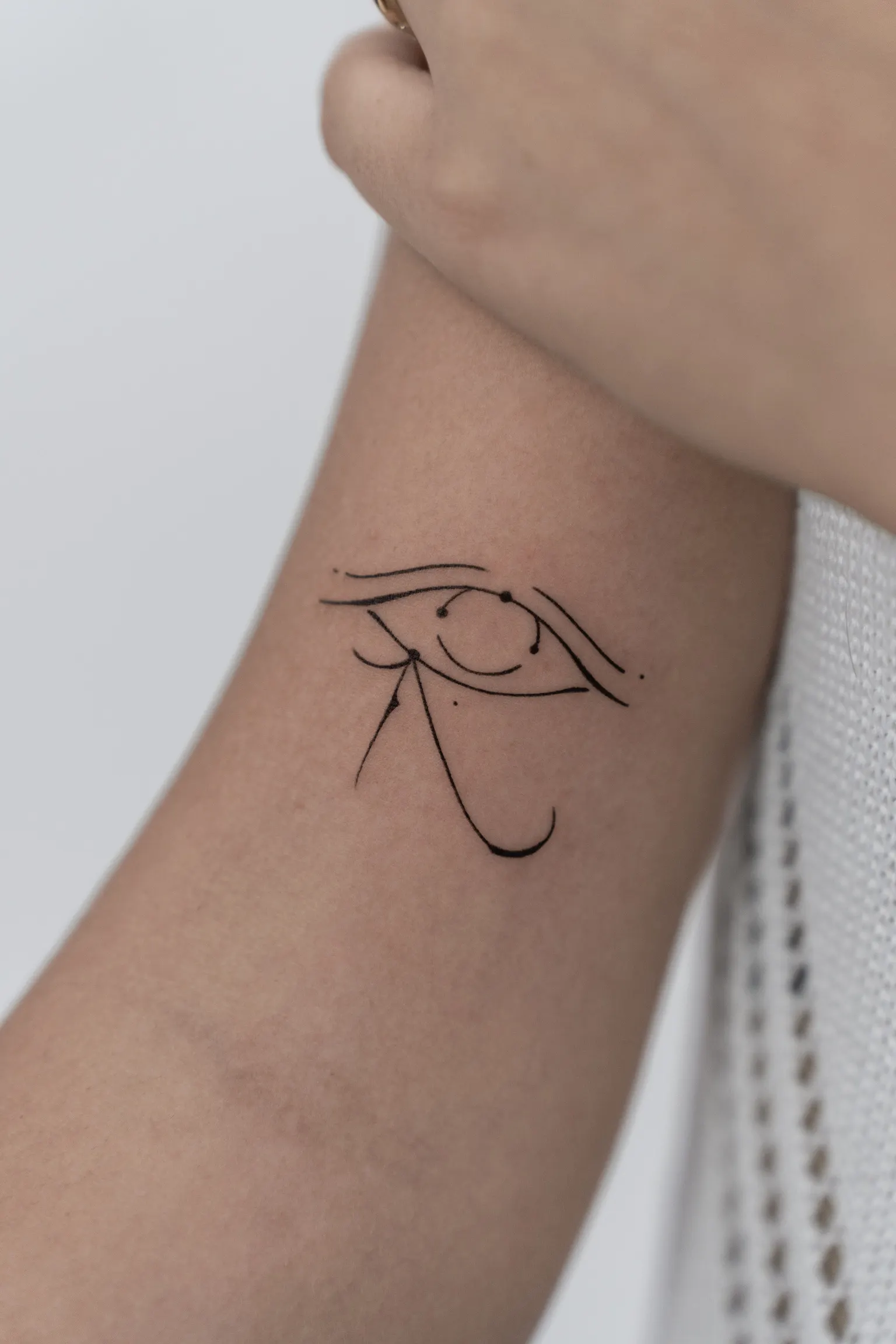 Fine-line eye symbol tattoo with flowing curved lines on the inner forearm.
