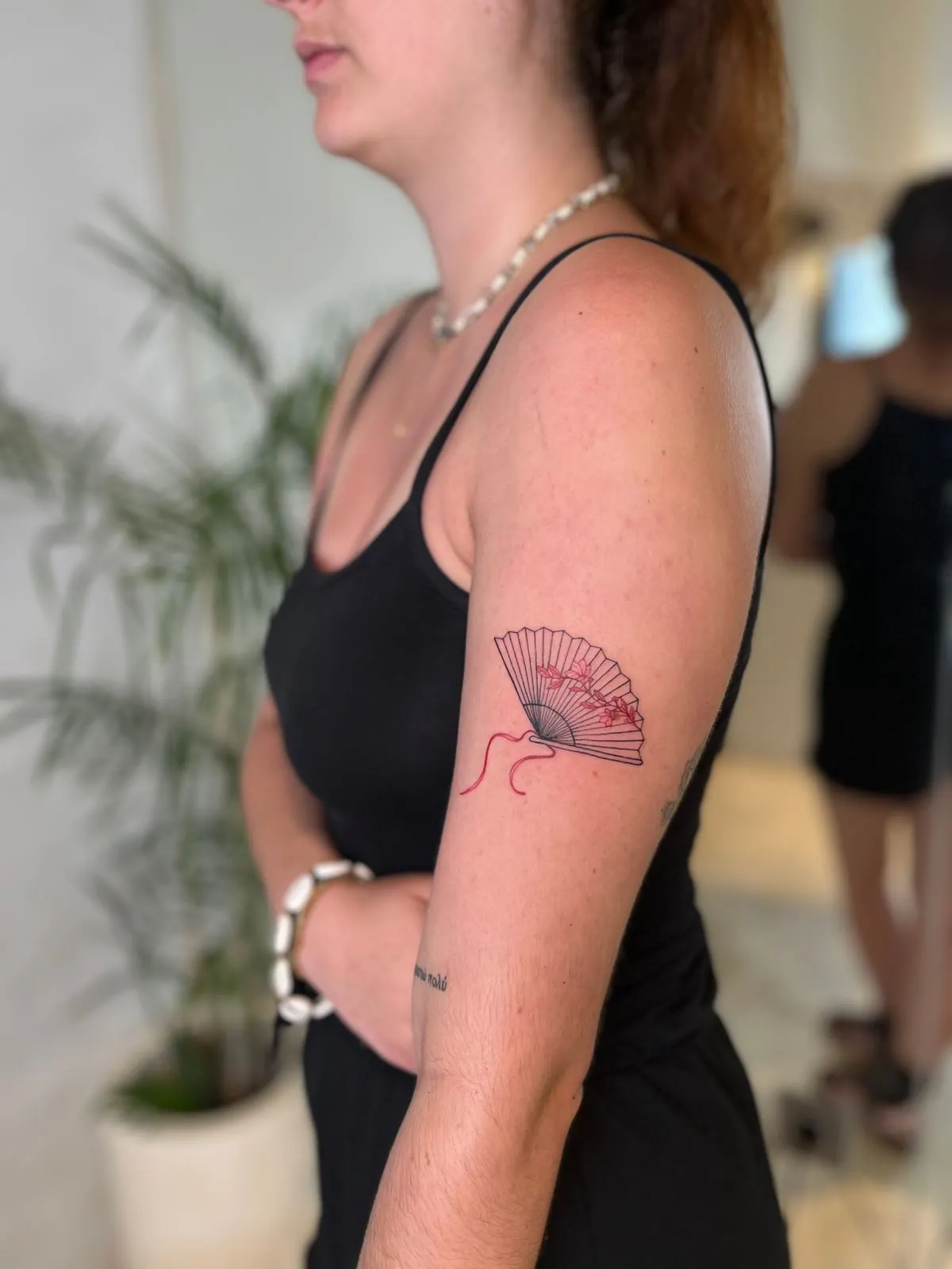 Tattoo of a folding fan with blossoms and a red ribbon on an upper arm.