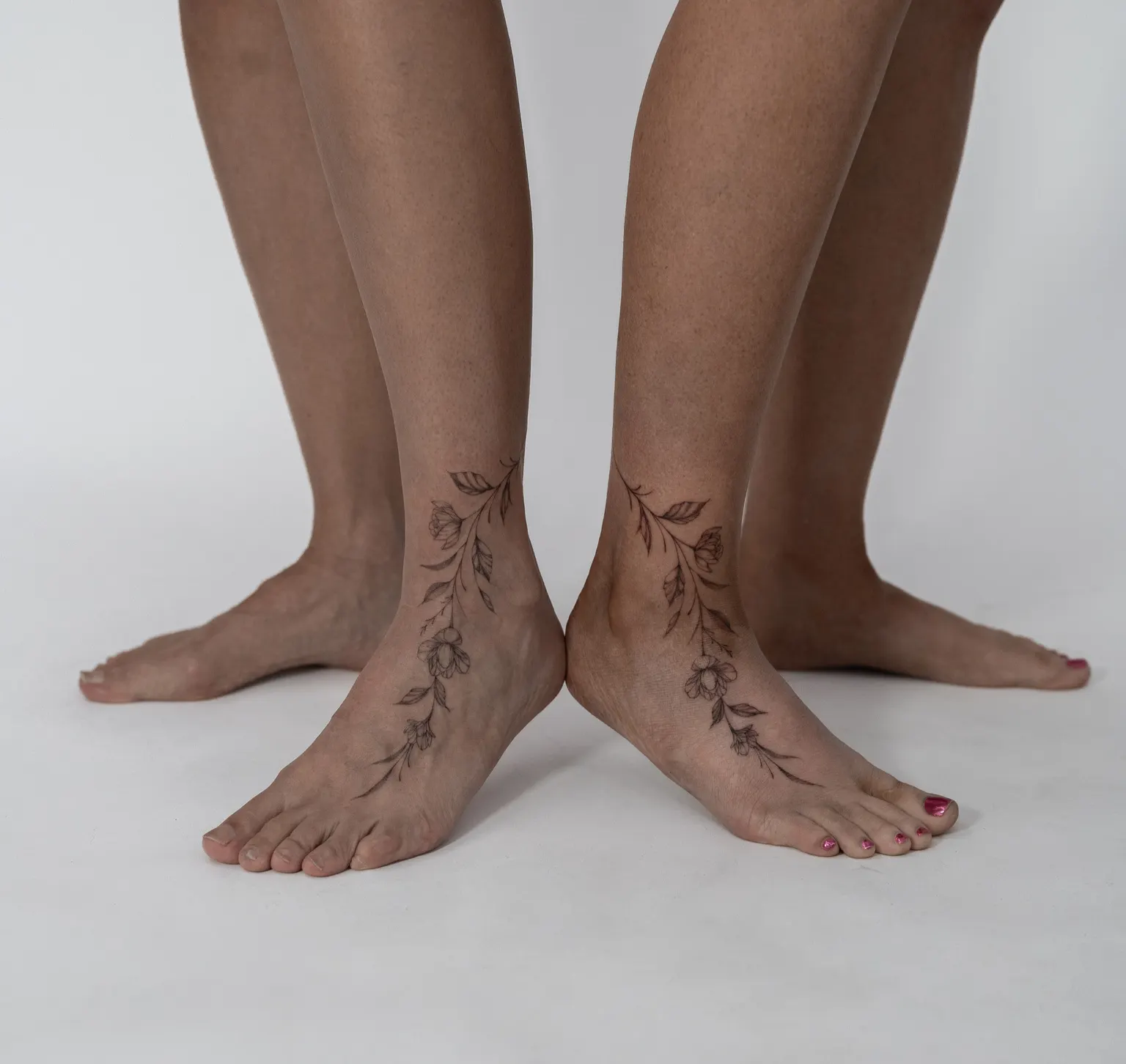 Fine-line floral vine tattoos running from both ankles onto the tops of the feet.