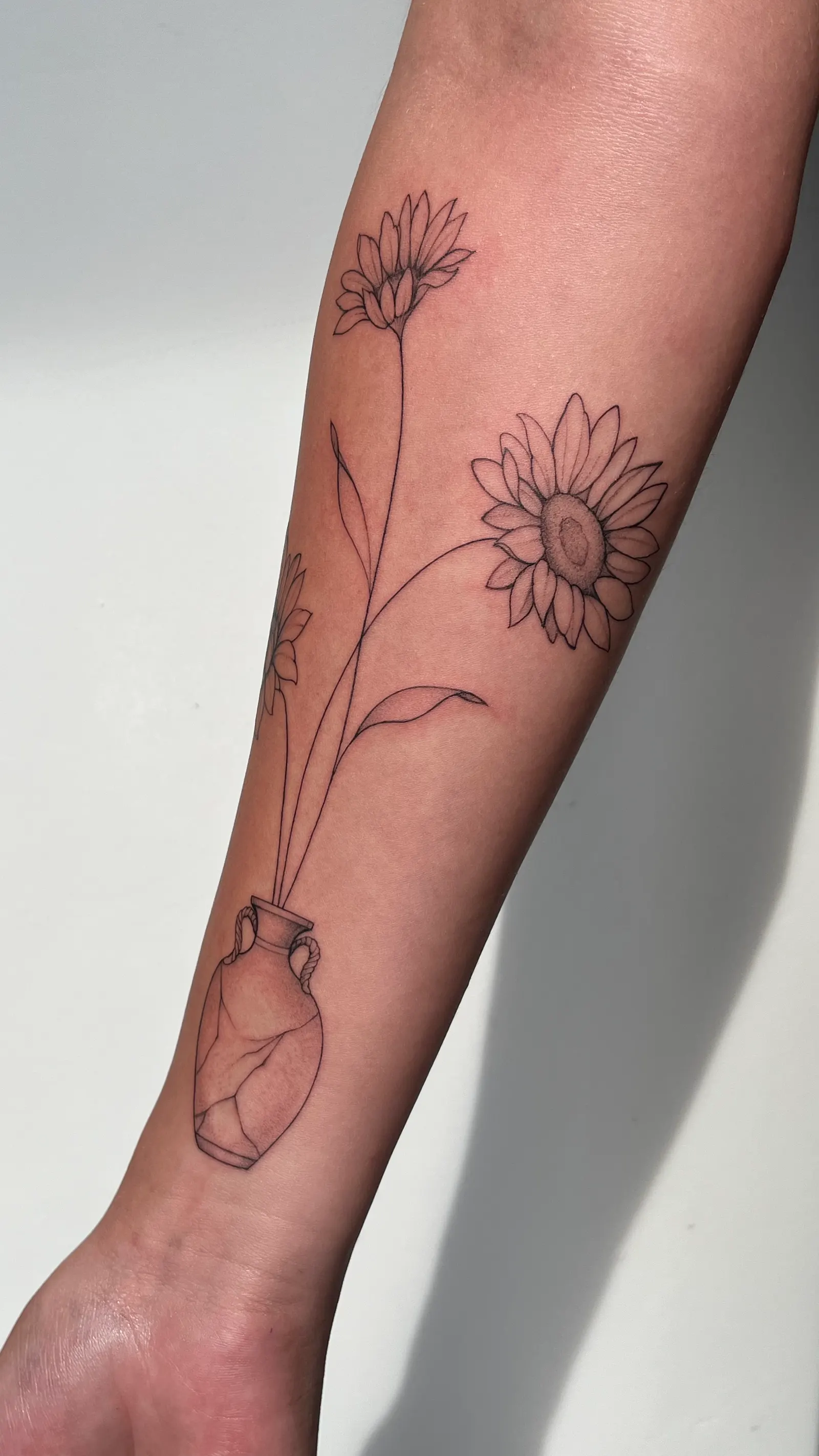 Fine-line tattoo of sunflowers and daisies in a vase running along a forearm.