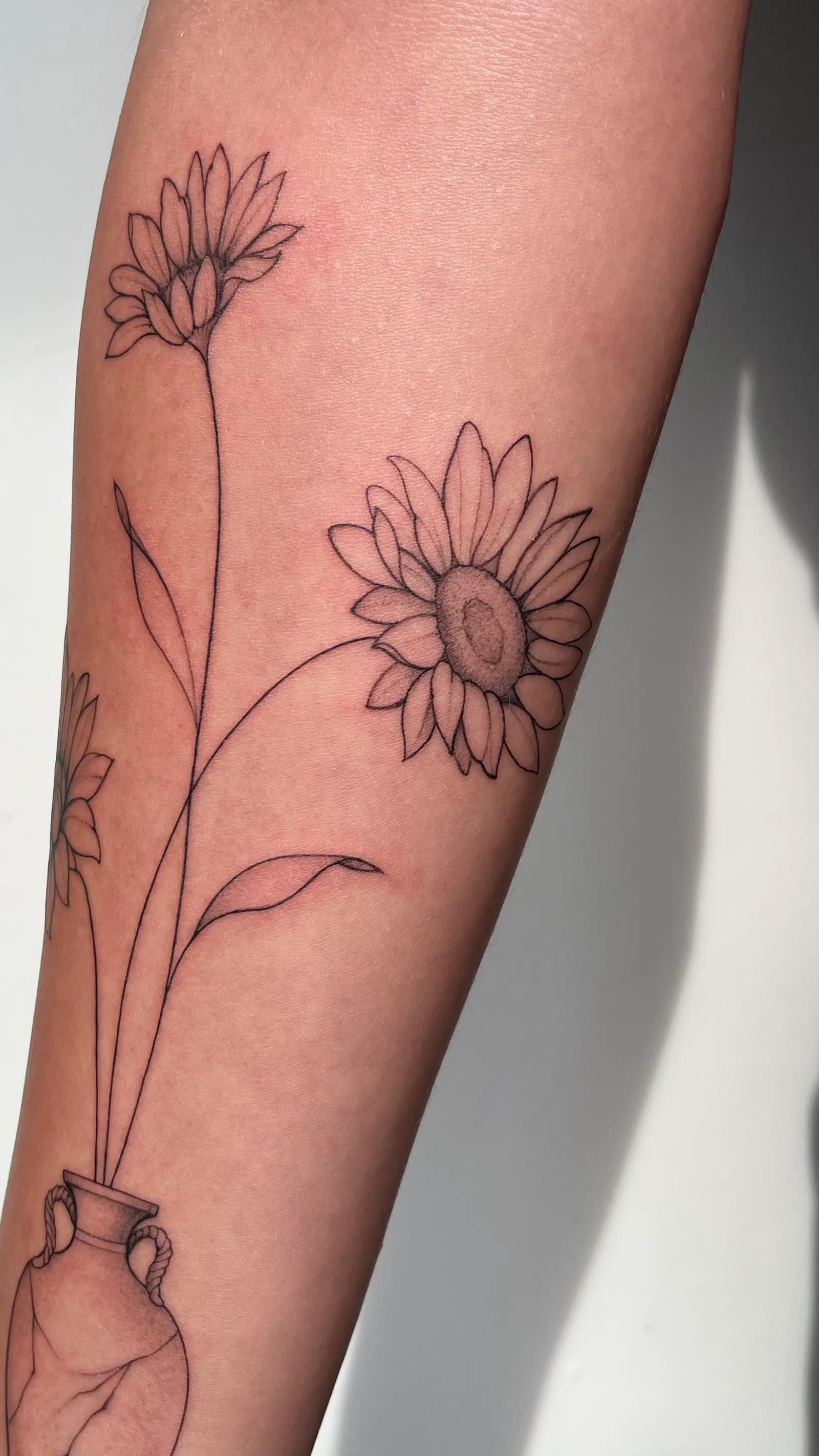 Close-up of a fine-line sunflower and daisy tattoo on a forearm.