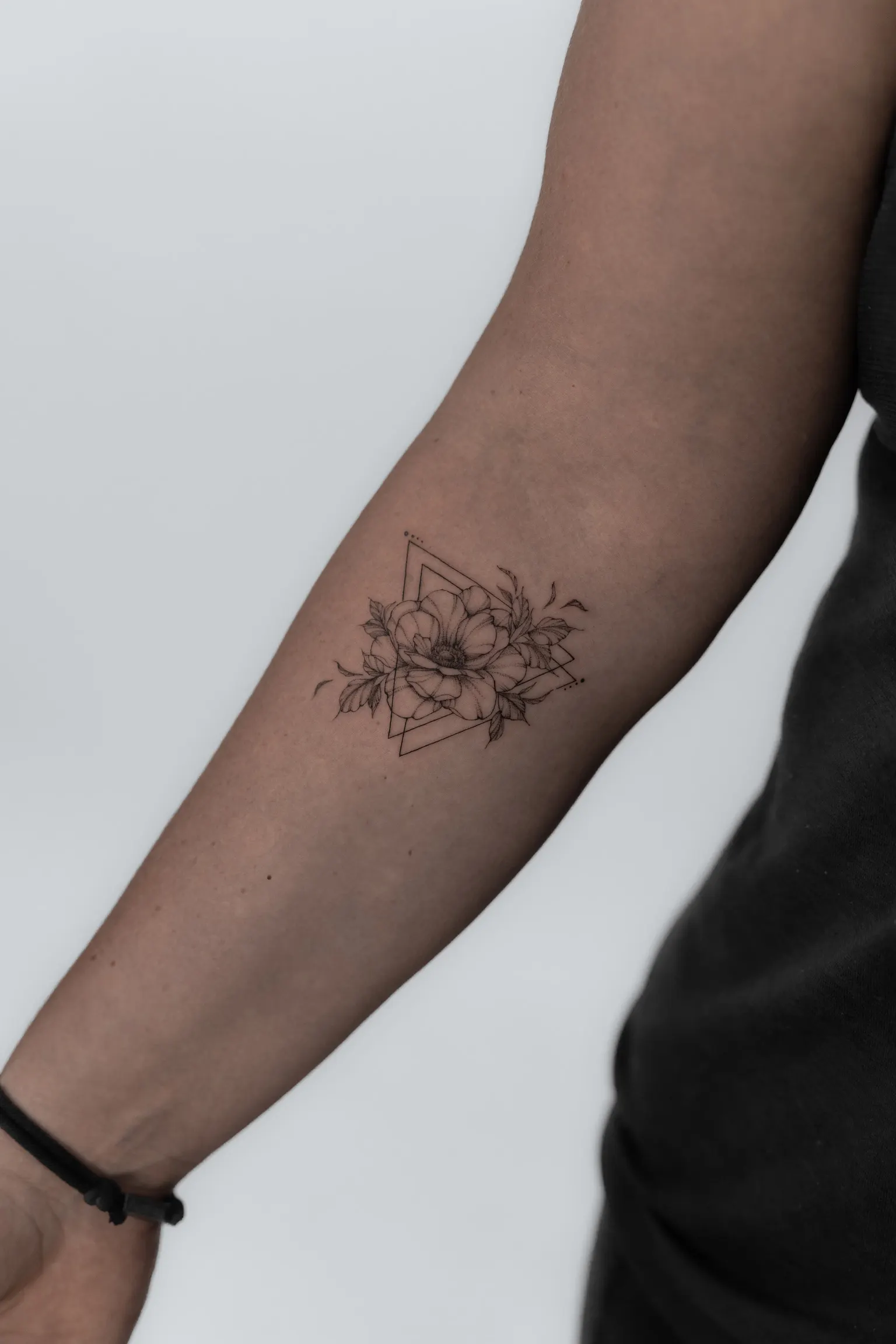Fine-line flower tattoo layered over geometric triangle outlines on a forearm.