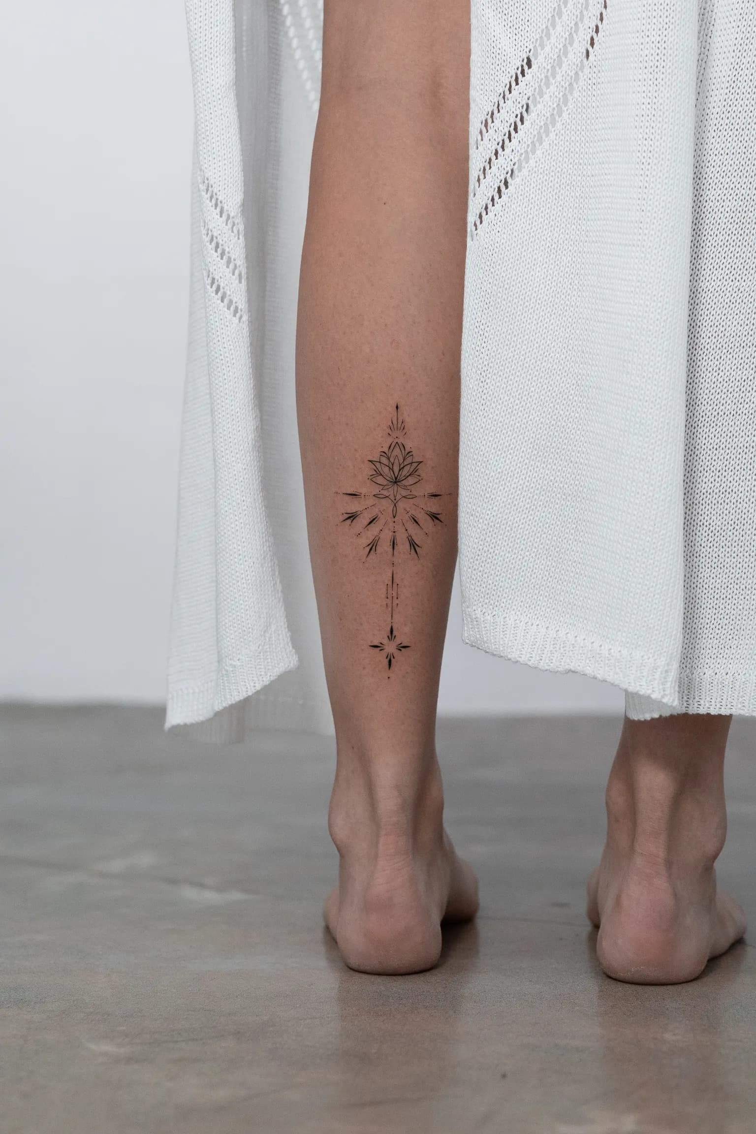 Fine-line ornamental lotus and arrow tattoo on the back of a lower leg.