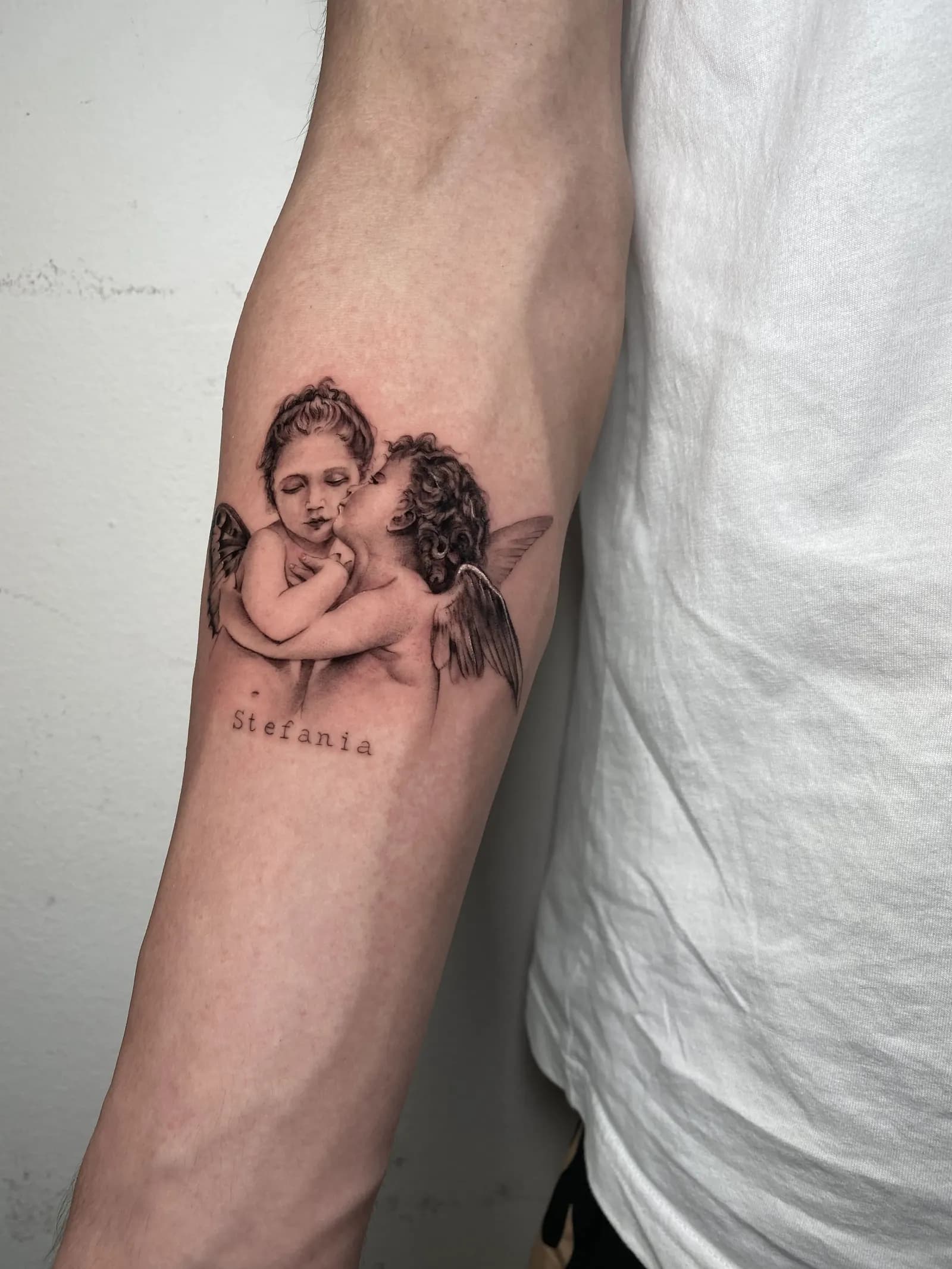 Black-and-grey tattoo of two cherubs above a name in small lettering on a forearm.