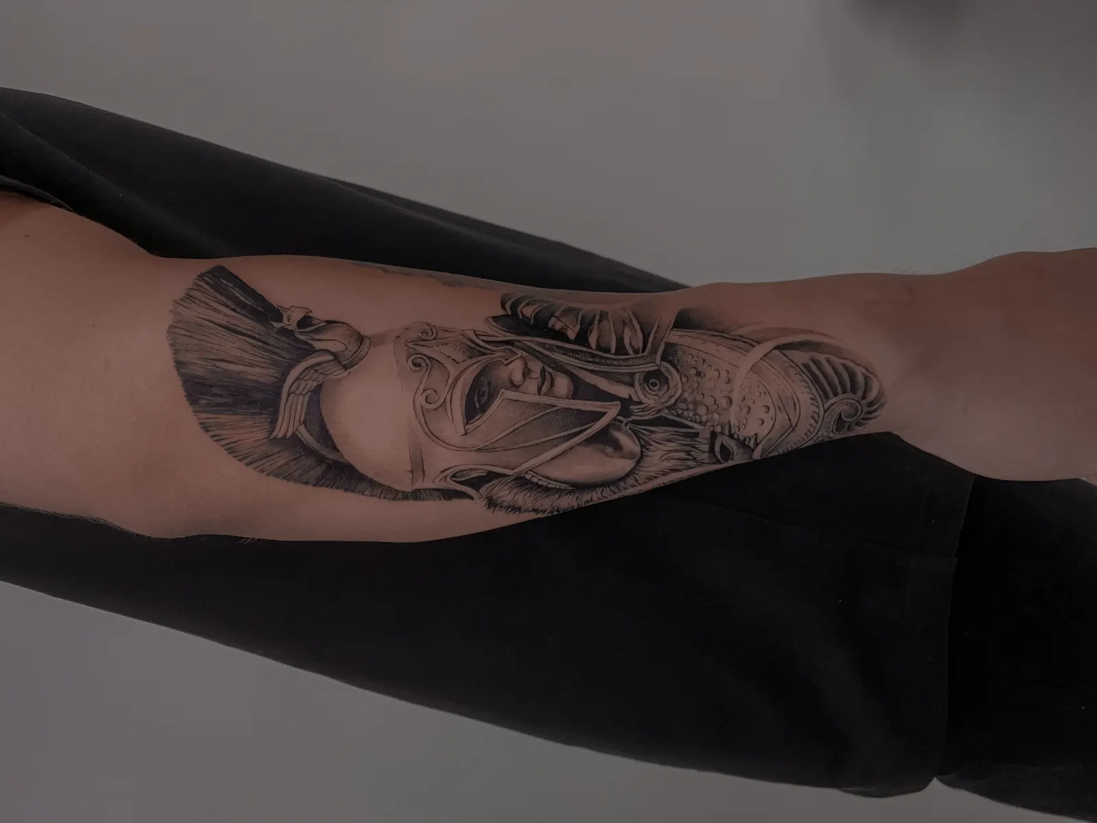 Black and grey portrait tattoo of a helmeted classical warrior on an inner upper arm.
