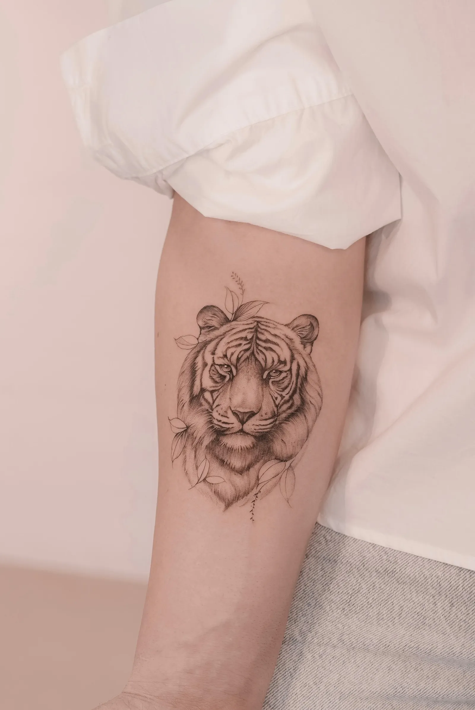 Black-and-grey tiger head framed by leaves tattooed on a forearm.
