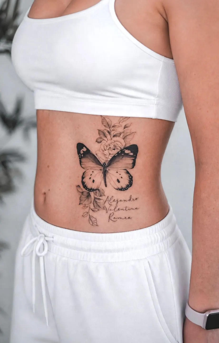 Black and grey butterfly and floral tattoo with three script names on a side torso.