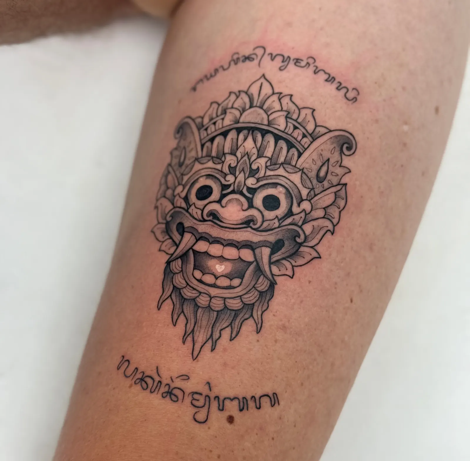 Black and grey ornamental mask tattoo with script lettering above and below, on a leg.