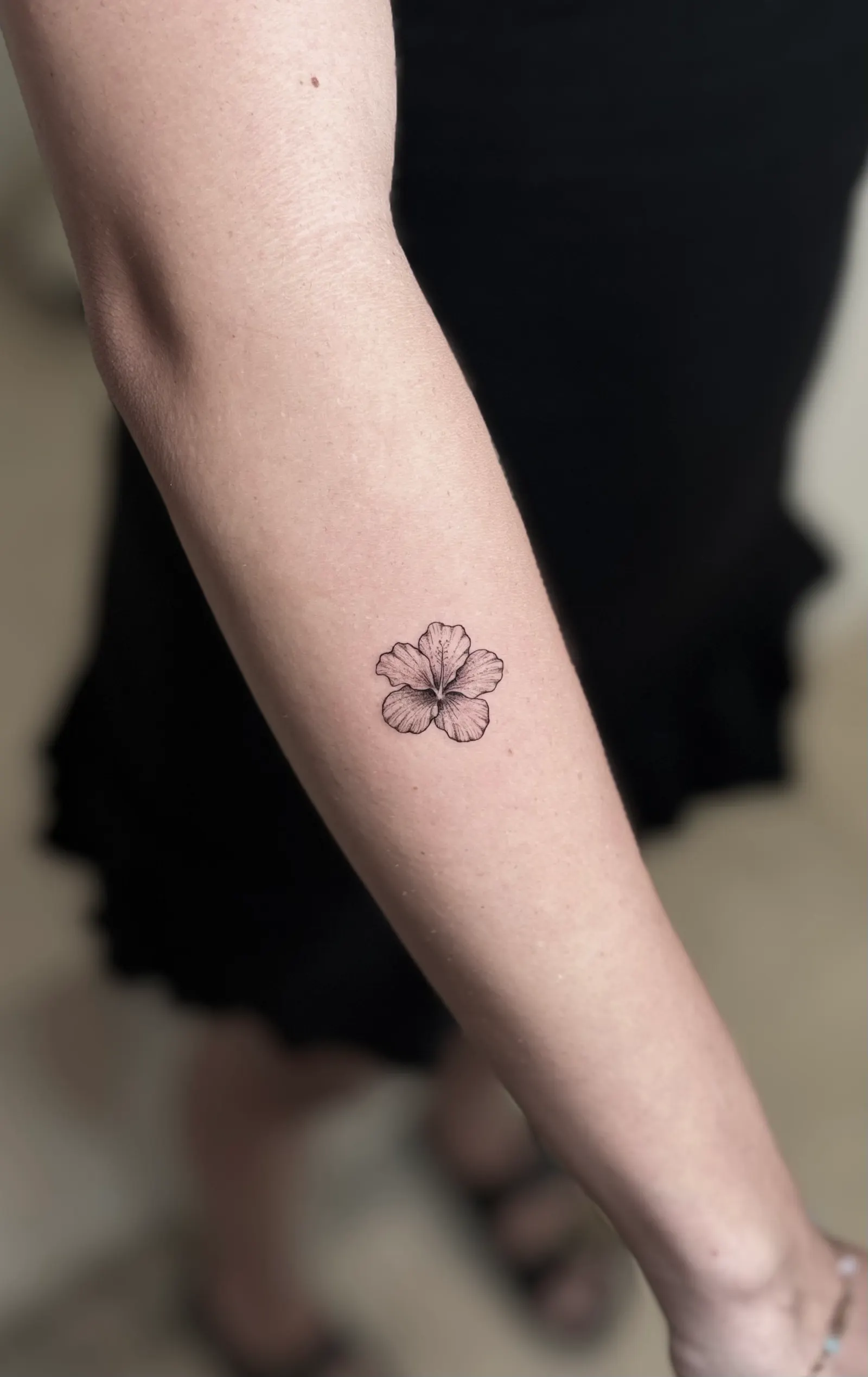 Small fine-line hibiscus flower tattoo on a forearm.