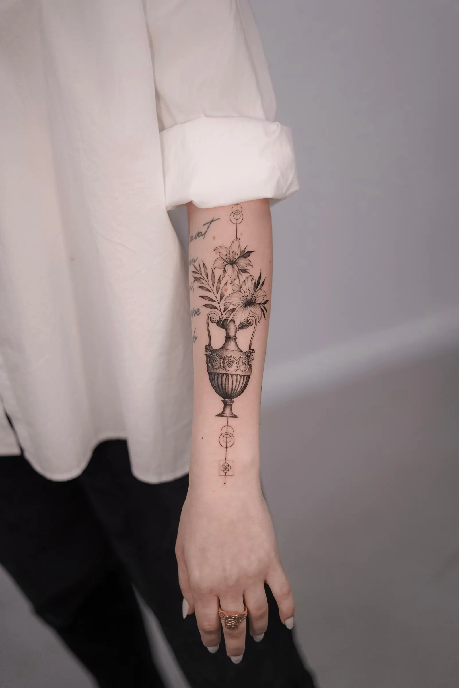 Ornate urn holding lilies with small geometric shapes above and below, on the forearm.