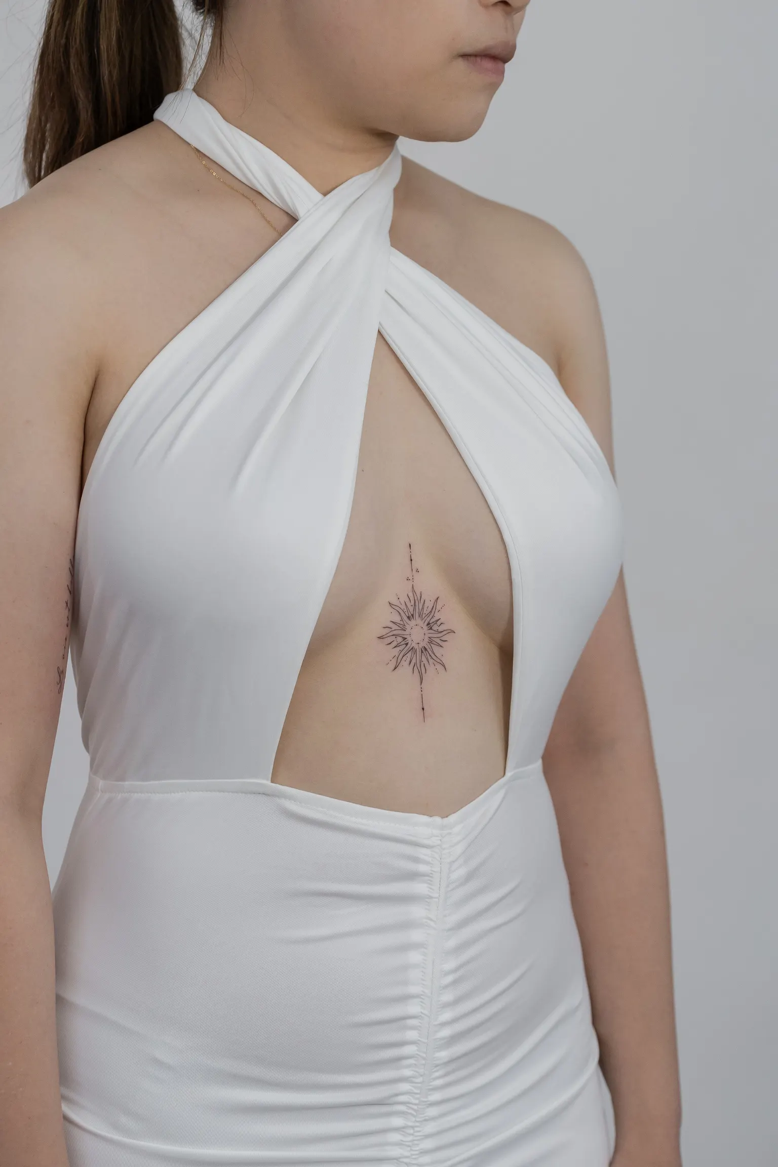 Fine-line sun and star burst tattoo on the sternum, framed by a white halter dress.
