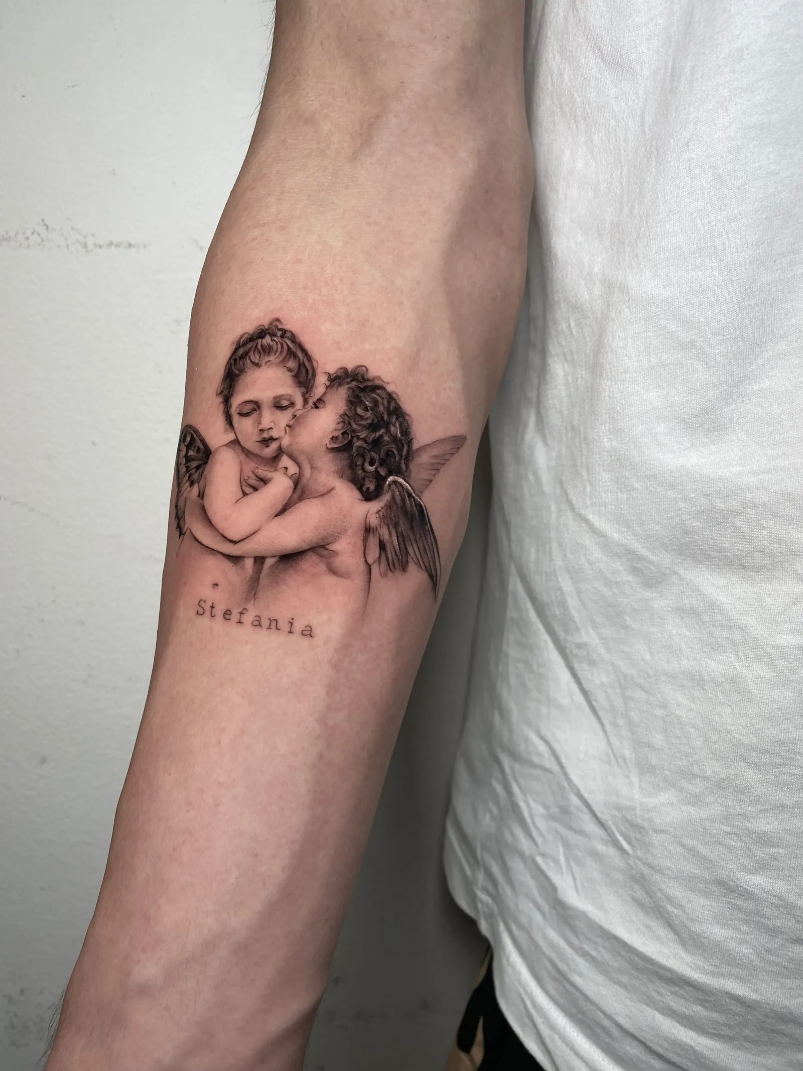 Black-and-grey tattoo of two cherubs above a name in small lettering on a forearm.