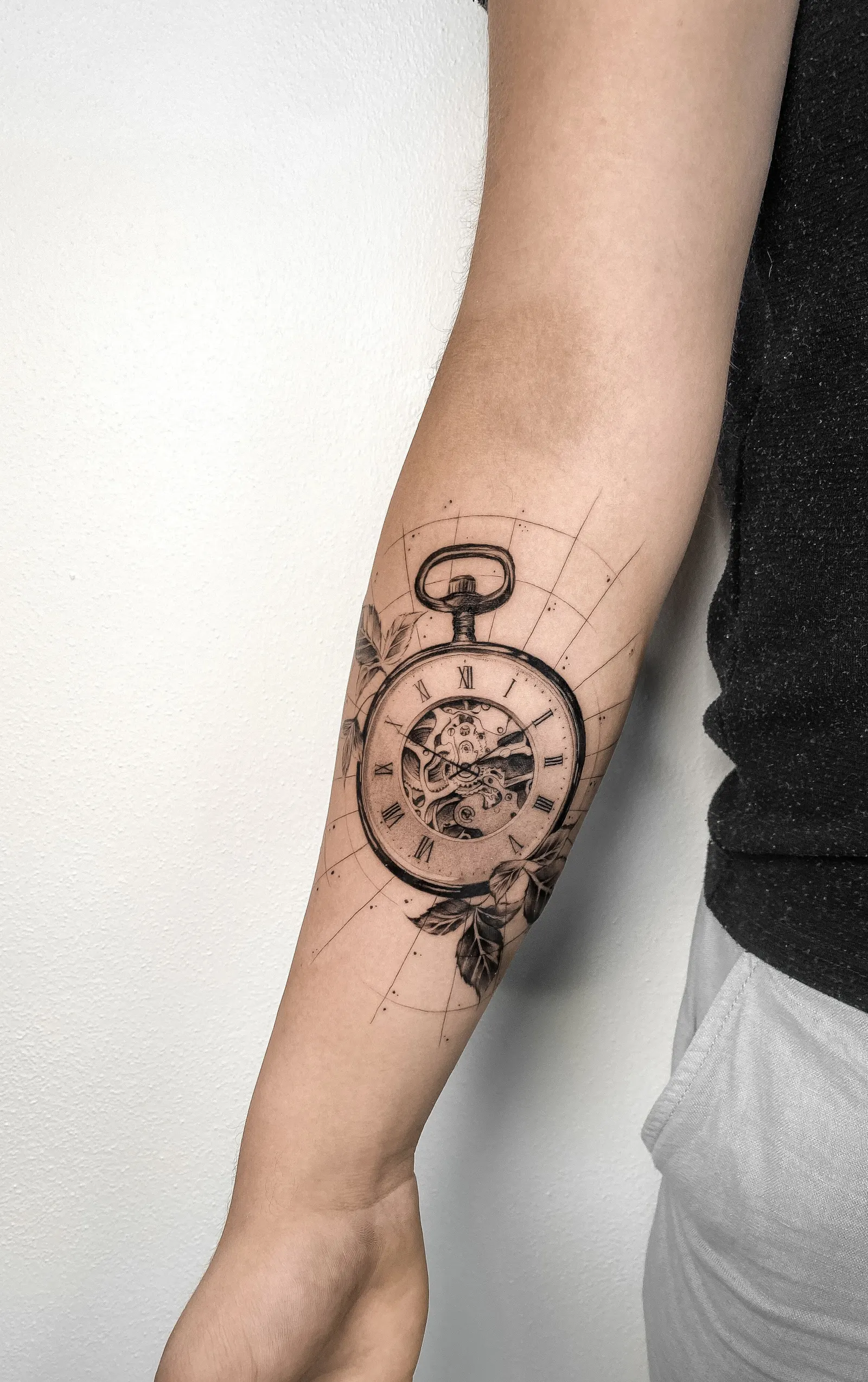 Black-and-grey pocket watch with leaves and thin arcs tattooed on a forearm.